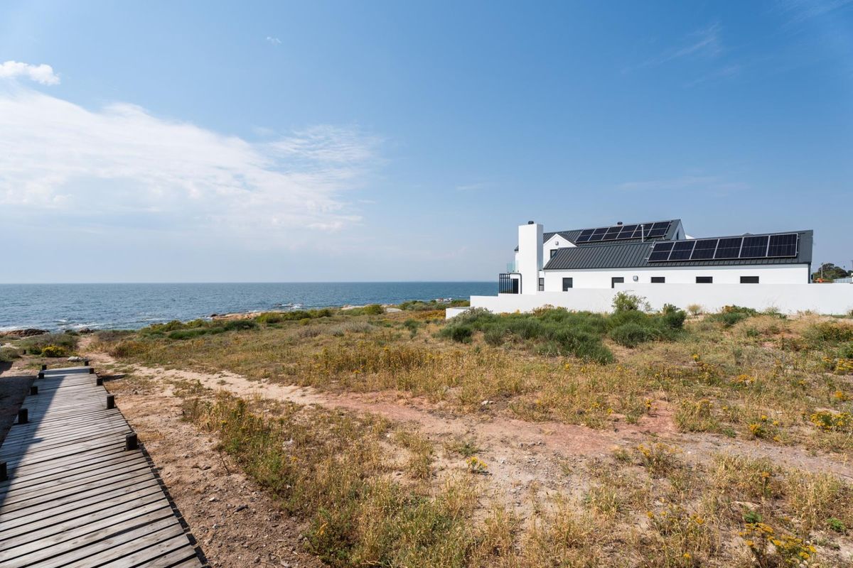 A scenic view of a modern coastal property featuring solar panels, a white exterior finish, and a wooden boardwalk leading towards the ocean.