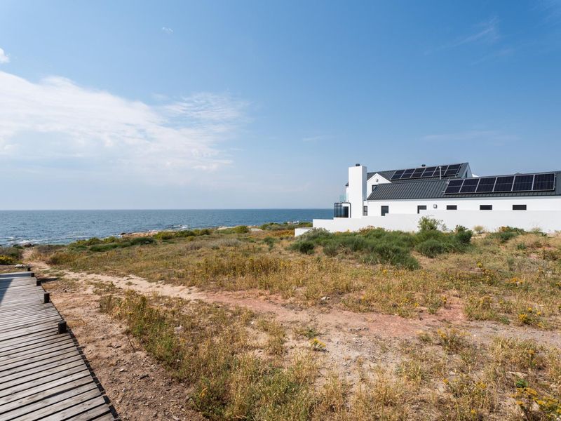 A scenic view of a modern coastal property featuring solar panels, a white exterior finish, and a wooden boardwalk leading towards the ocean.