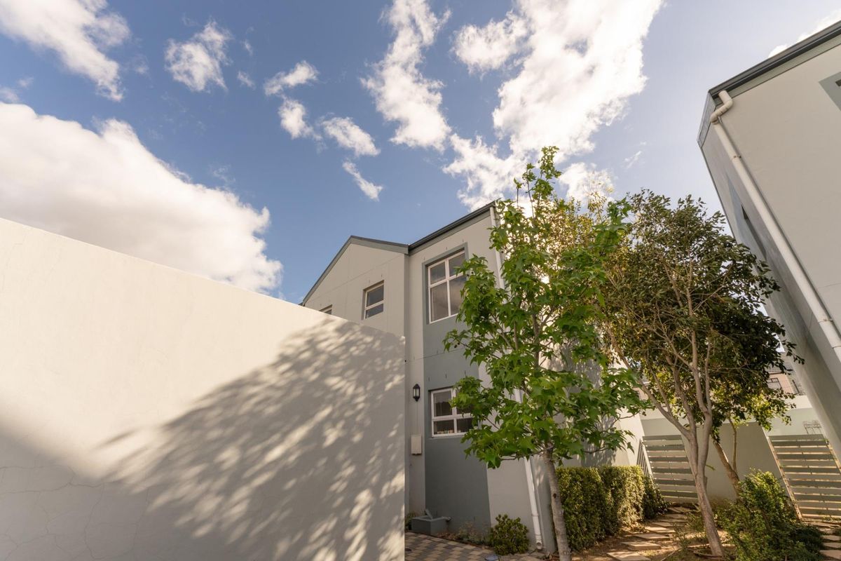 A view of the rear exterior of the house featuring a paved walkway, boundary wall, and established trees.