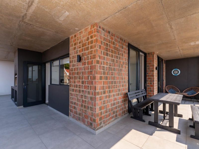 Spacious covered balcony featuring exposed concrete ceilings, brick architectural detailing, tiled flooring, and a built-in braai area.