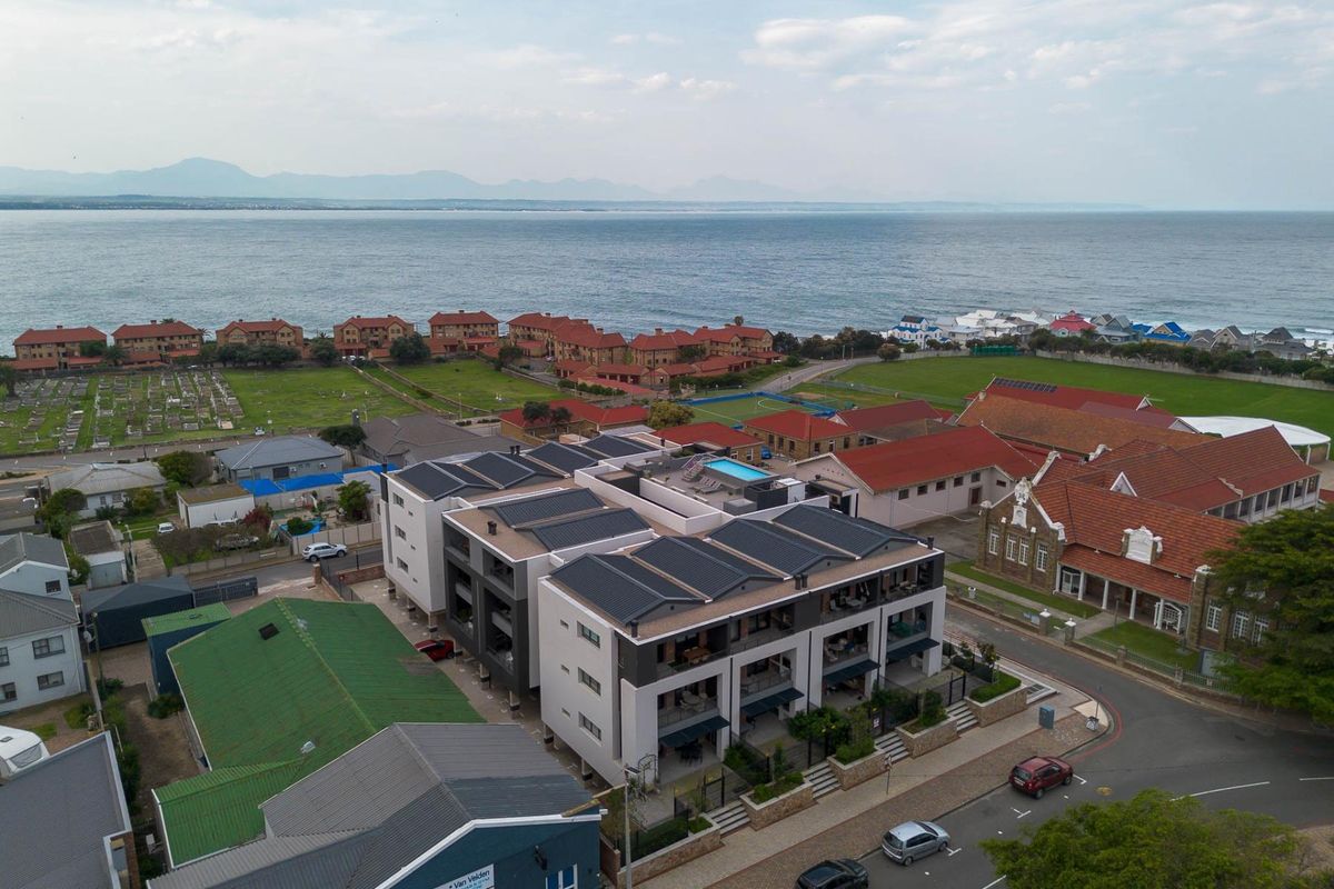 Aerial view of a modern residential complex featuring rooftop solar panels, a swimming pool, and balconies, situated near the coastline.