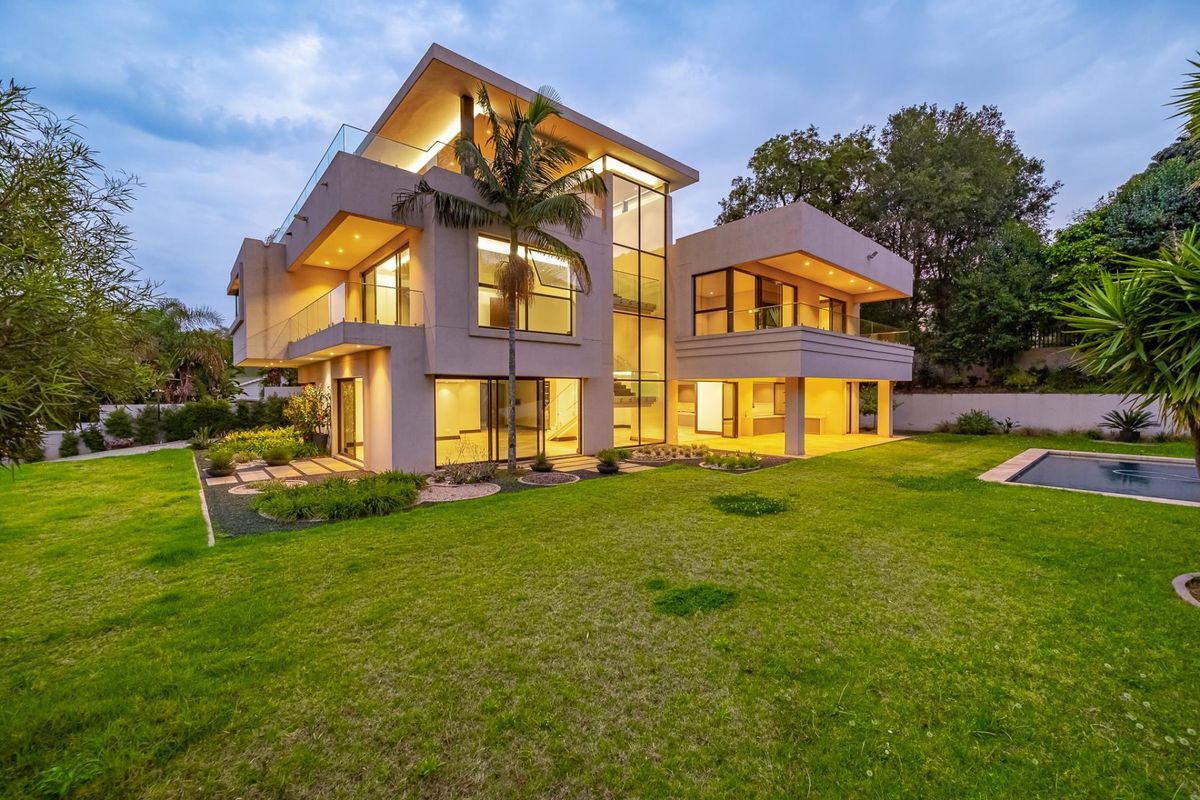 Modern multi-story luxury residence featuring expansive glass facades, integrated lighting, a covered patio, and a well-maintained garden with a swimming pool.
