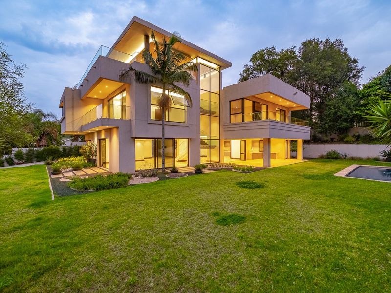 Modern multi-story luxury residence featuring expansive glass facades, integrated lighting, a covered patio, and a well-maintained garden with a swimming pool.