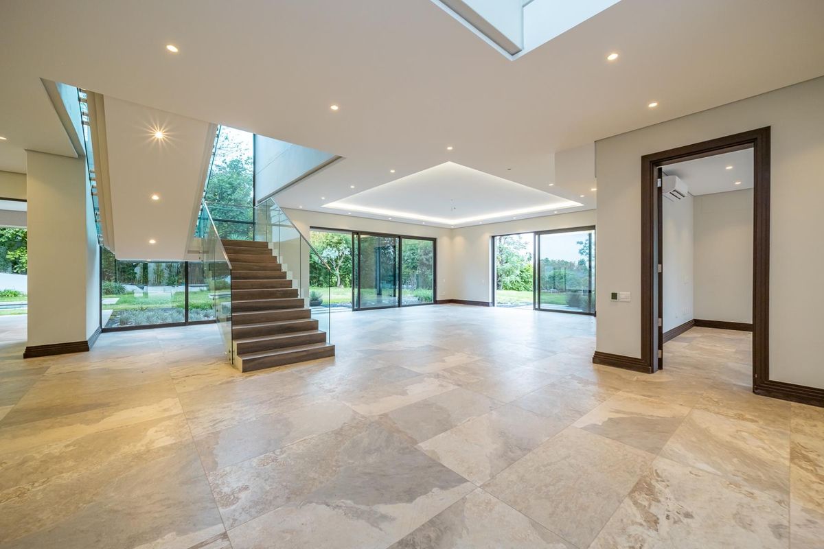 Spacious open-plan living area featuring large format stone tiles, a modern staircase with glass balustrades, recessed lighting, and expansive sliding doors leading to the garden.