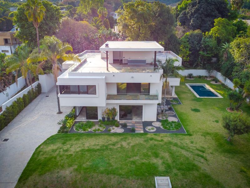 Aerial view of a modern multi-level residence featuring a flat roof, glass balustrades, a swimming pool, and a spacious landscaped garden.