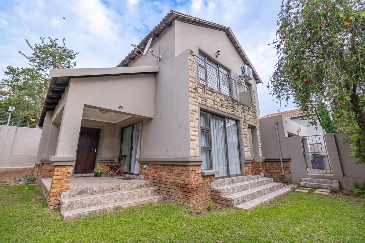 Rear view of a double-story house featuring stone cladding, a tiled patio, a balcony, and a well-maintained garden area.