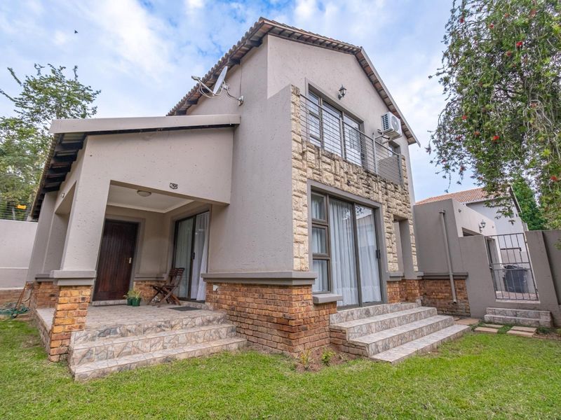 Rear view of a double-story house featuring stone cladding, a tiled patio, a balcony, and a well-maintained garden area.