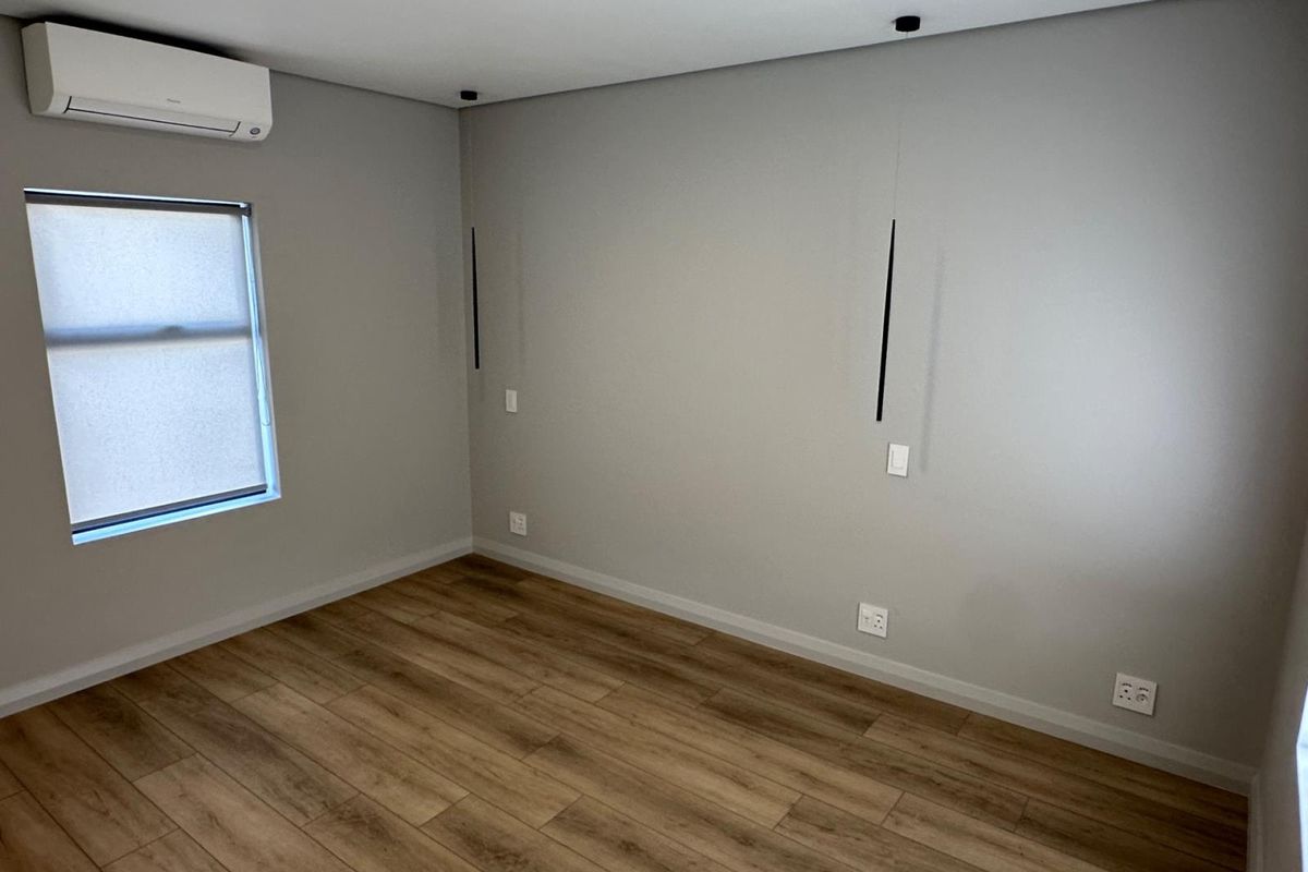 A clean, modern, and empty bedroom featuring laminate wood flooring, neutral grey walls, a split-system air conditioner, and contemporary wall-mounted lighting.