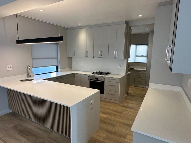 Modern kitchen featuring light grey cabinetry, stone countertops, a central island, and integrated appliances, with a separate scullery area visible in the background.