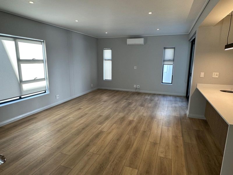 A bright and modern open-plan living area featuring wood-look laminate flooring, recessed lighting, a wall-mounted air conditioner, and a sleek kitchen counter.