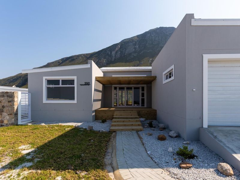 Modern single-story home exterior featuring a timber deck entrance, paved walkway, and integrated garage, set against a scenic mountain backdrop.