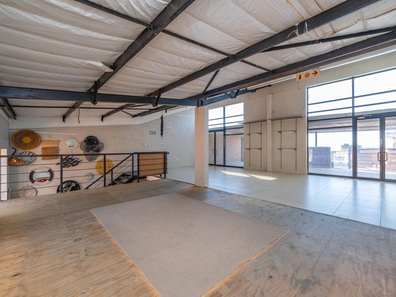 Spacious open-plan retail unit featuring high ceilings with exposed steel trusses, large glass windows for natural light, and a mezzanine level with plywood flooring.