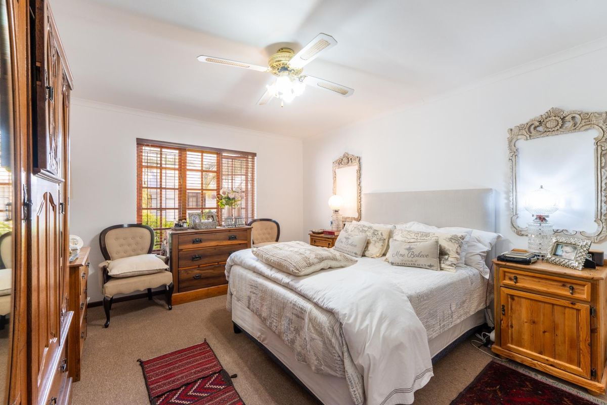 A well-lit bedroom featuring carpeted flooring, a ceiling fan, and wooden window blinds, offering a comfortable and functional living space.