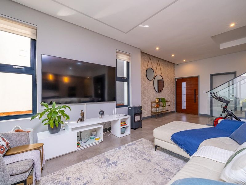 Modern open-plan living area featuring tiled floors, recessed lighting, a glass staircase balustrade, and a solid wood entrance door.