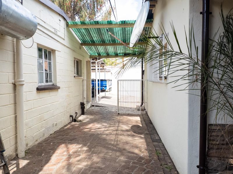 A narrow rear exterior walkway featuring brick paving, a corrugated plastic covered section, and a metal security gate.