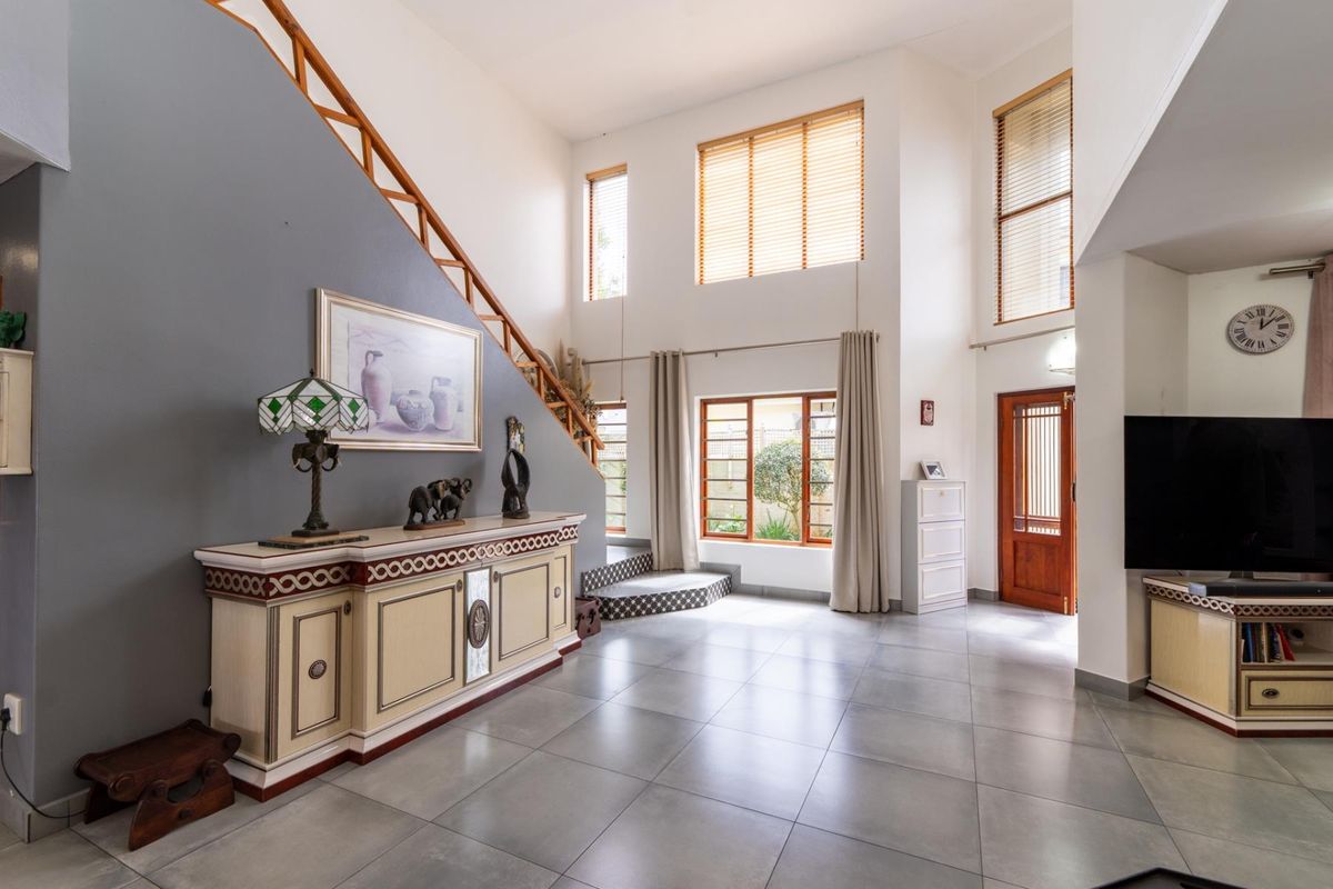 Spacious double-volume living area featuring large windows, tiled flooring, and a staircase with a wooden handrail, creating a bright and open atmosphere.