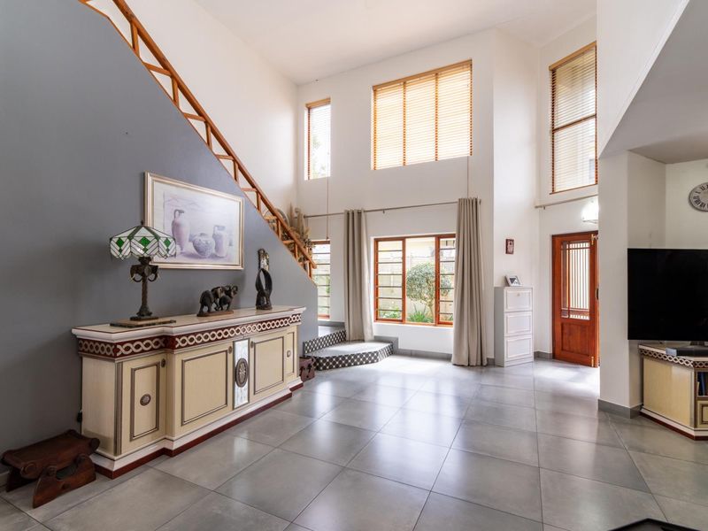 Spacious double-volume living area featuring large windows, tiled flooring, and a staircase with a wooden handrail, creating a bright and open atmosphere.
