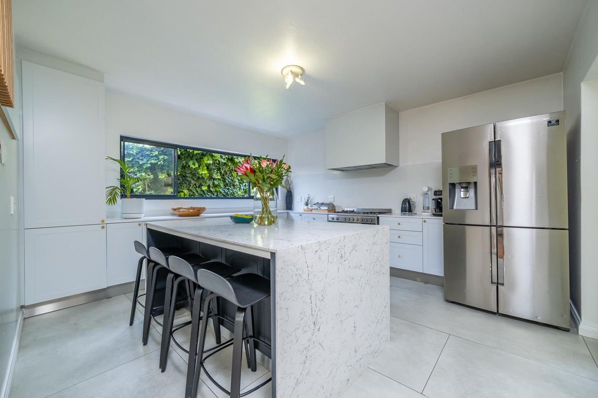 Modern kitchen featuring a large stone-topped island, sleek white cabinetry, integrated appliances, and large windows overlooking the garden.