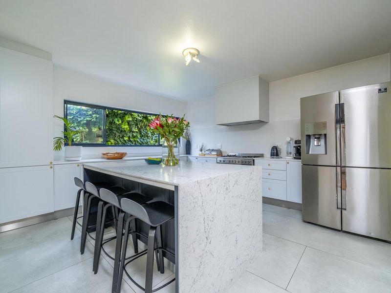 Modern kitchen featuring a large stone-topped island, sleek white cabinetry, integrated appliances, and large windows overlooking the garden.