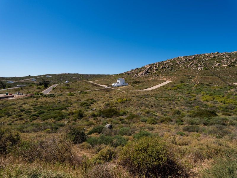 An aerial view of a large, undeveloped residential land plot featuring natural vegetation, rocky terrain, and an existing paved access road.