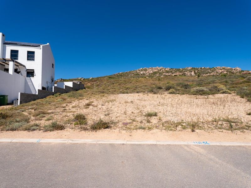 A vacant residential plot of land situated next to an existing white building, featuring natural terrain and street access.