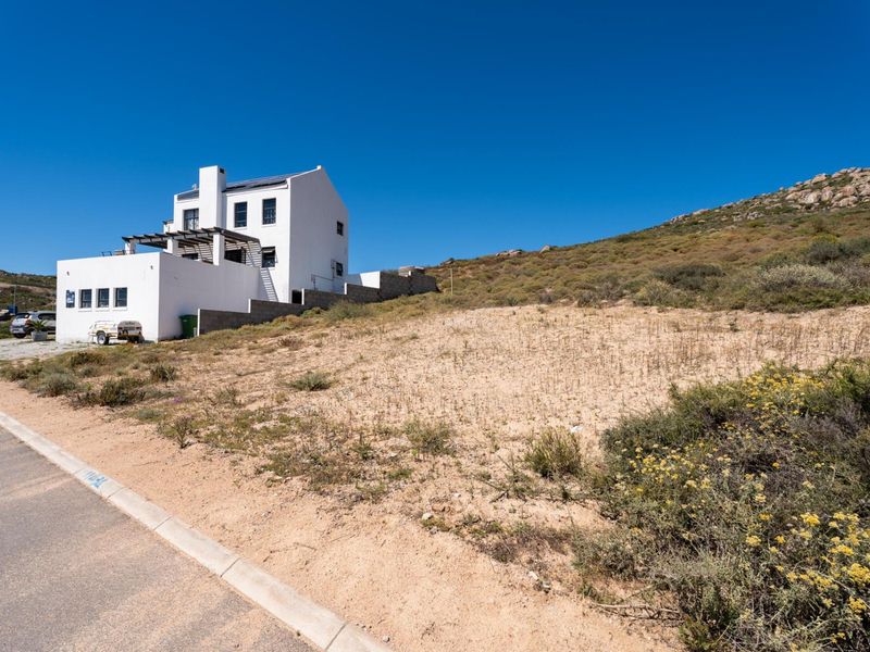 A vacant residential plot situated next to an existing white multi-story dwelling, featuring natural vegetation and access from a paved street.
