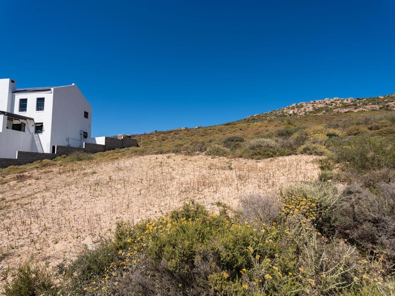 A view of a vacant residential plot featuring natural sloped terrain and native vegetation, adjacent to an existing white residential structure.