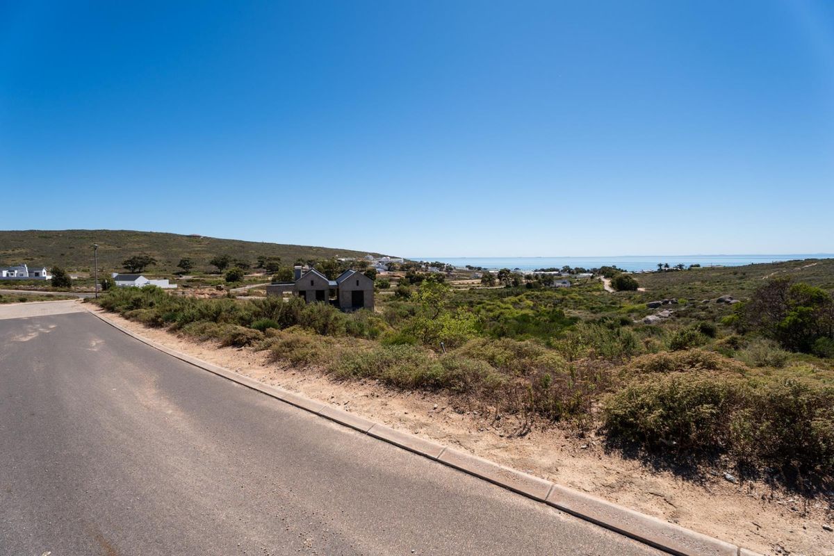 A street-level view of a vacant residential plot featuring natural coastal vegetation, paved road access, and distant ocean views.