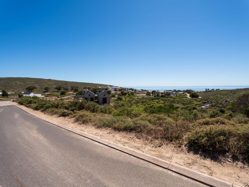 A street-level view of a vacant residential plot featuring natural coastal vegetation, paved road access, and distant ocean views.