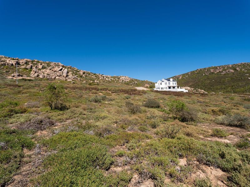 An elevated view of a large, undeveloped residential plot featuring natural rocky terrain and native fynbos vegetation with a distant house visible.