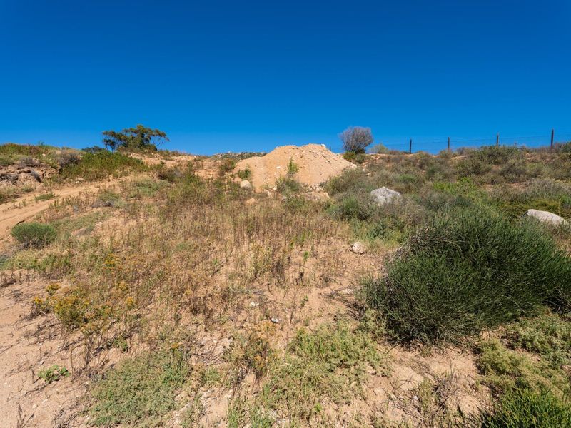 A view of the vacant residential land featuring natural vegetation, a dirt mound, and a perimeter wire fence against a clear blue sky.