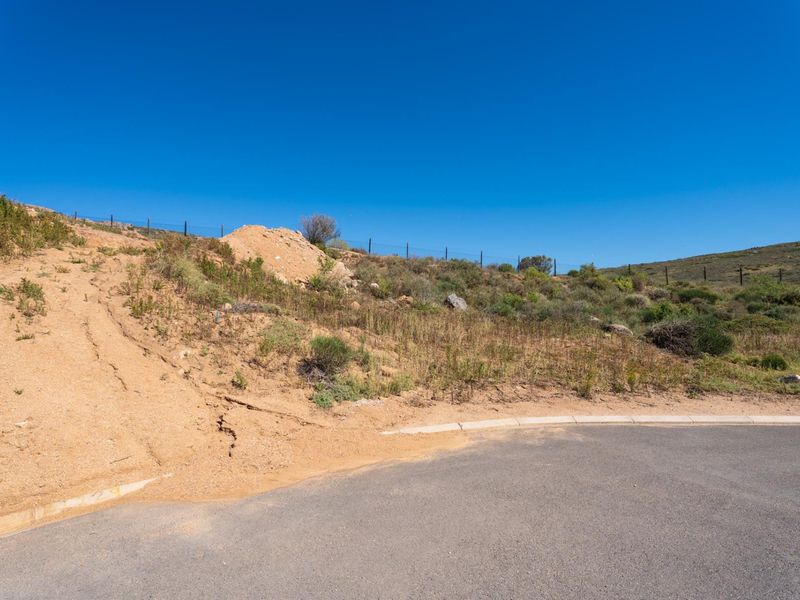 A vacant residential plot featuring natural sloping terrain, perimeter fencing, and direct access to an asphalt road with concrete curbing.