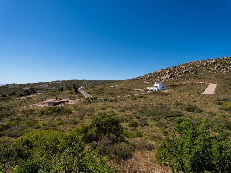 An aerial view of a residential development site featuring natural landscape, paved access roads, and existing residential structures in the distance.