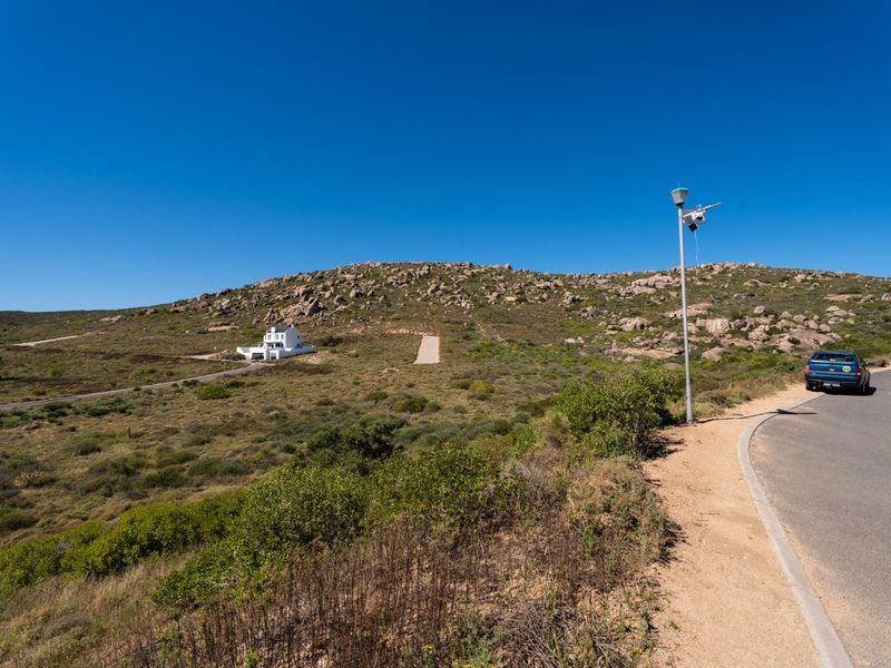 A wide-angle view of a residential landscape featuring a paved road, street lighting, and a distant house set against a rocky hillside.