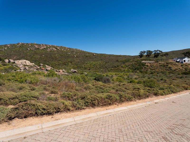 A view of a vacant residential plot from the paved street, featuring natural fynbos vegetation and rocky hillside terrain.