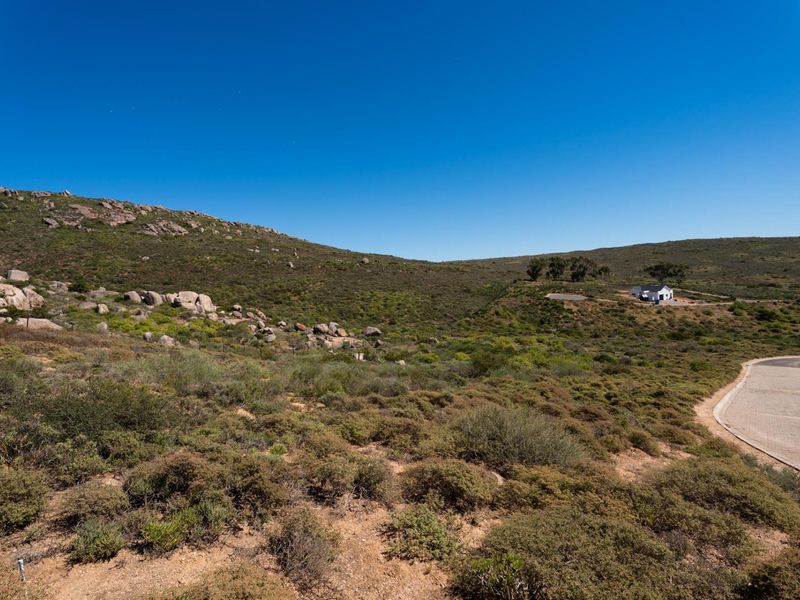A wide-angle view of the vacant residential land, featuring natural rocky terrain, native vegetation, and an adjacent paved access road.