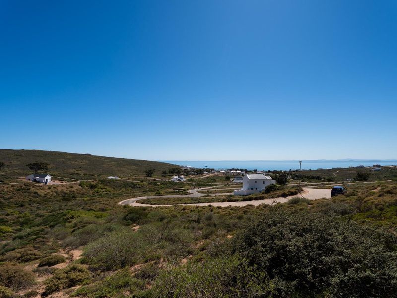 An elevated aerial view of a residential development area featuring winding paved roads, natural coastal vegetation, and distant ocean views.