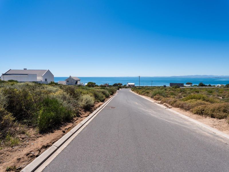 A clear street view of a paved residential road leading towards the ocean, showcasing the coastal location and surrounding vacant land.
