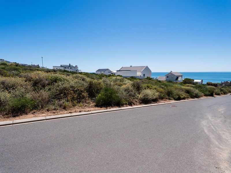 A street-level view of a vacant residential plot featuring natural coastal vegetation, paved road access, and distant ocean views.