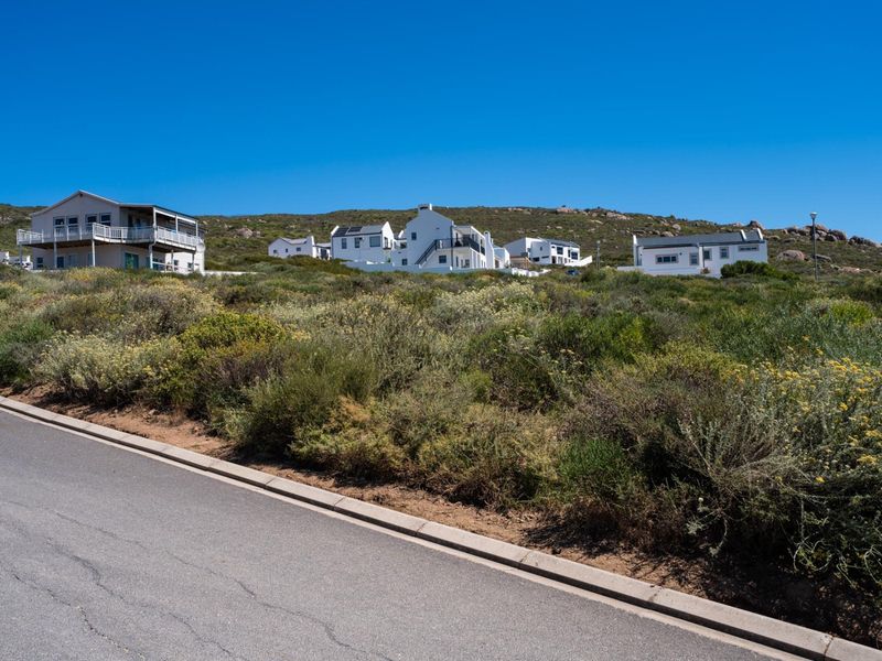 A street-level view of a vacant residential plot featuring natural coastal vegetation, with established neighboring homes visible in the background.