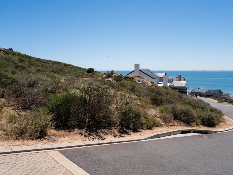 A scenic view of a vacant residential plot featuring natural coastal vegetation, overlooking the ocean and nearby residential properties.
