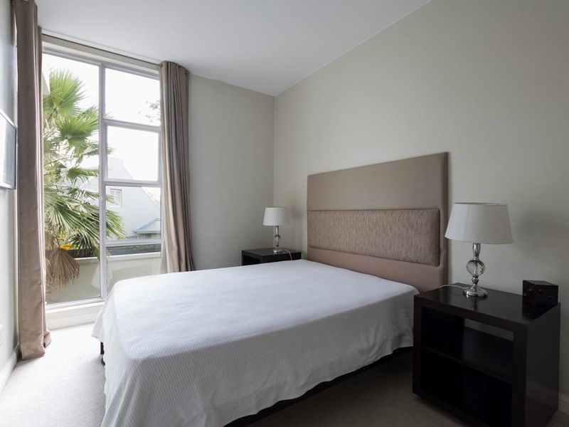 A well-lit bedroom featuring neutral walls, carpeted flooring, and a large floor-to-ceiling window providing natural light and a view of the outdoors.