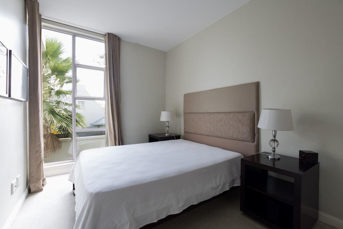 A well-lit bedroom featuring neutral walls, carpeted flooring, and a large floor-to-ceiling window providing natural light and a view of the outdoors.