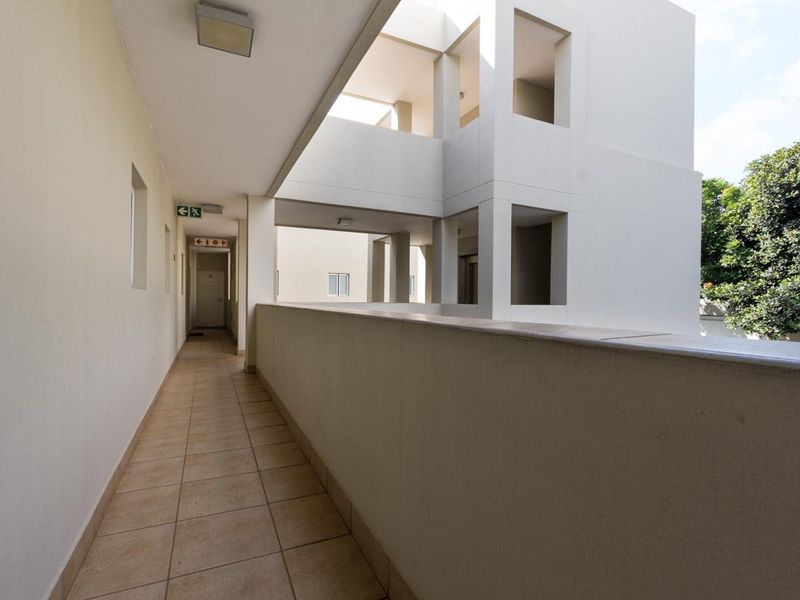 A clean, modern outdoor apartment hallway featuring tiled flooring, a protective balustrade, and clear emergency exit signage.