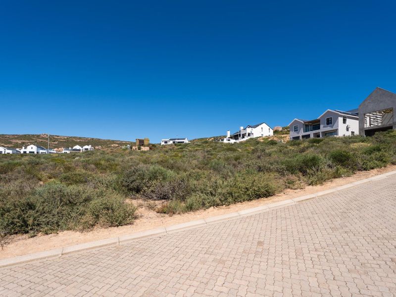 A view of a vacant residential plot from the paved street, featuring natural coastal vegetation and surrounding established homes.