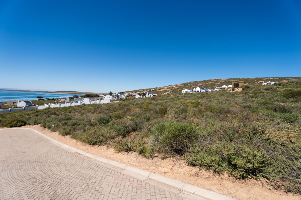 A view of a vacant residential plot from the paved street, featuring natural coastal vegetation and surrounding neighborhood homes.