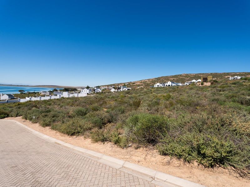 A view of a vacant residential plot from the paved street, featuring natural coastal vegetation and surrounding neighborhood homes.