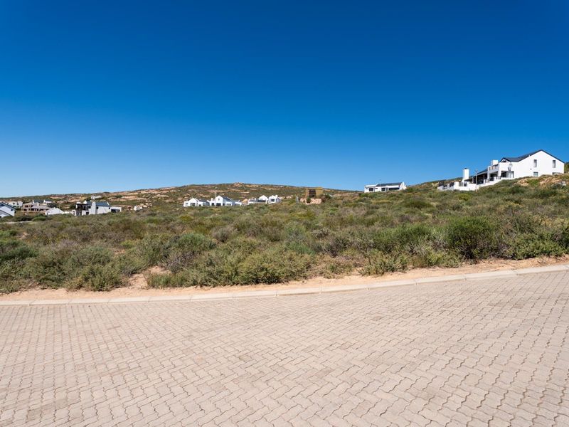 A view of a vacant residential plot featuring a paved driveway and natural coastal vegetation in a developed estate setting.