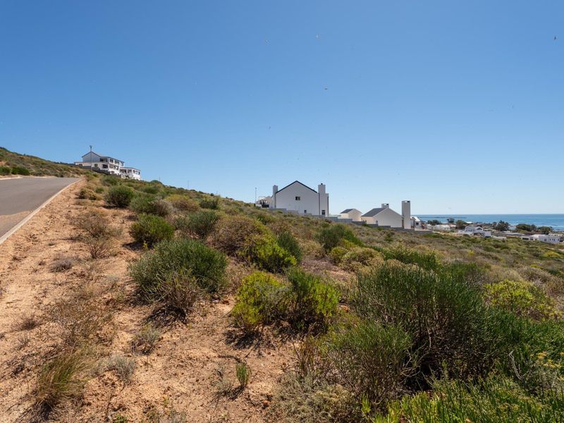 A view of a paved road bordering a vacant residential plot with coastal views and surrounding neighborhood homes in the distance.