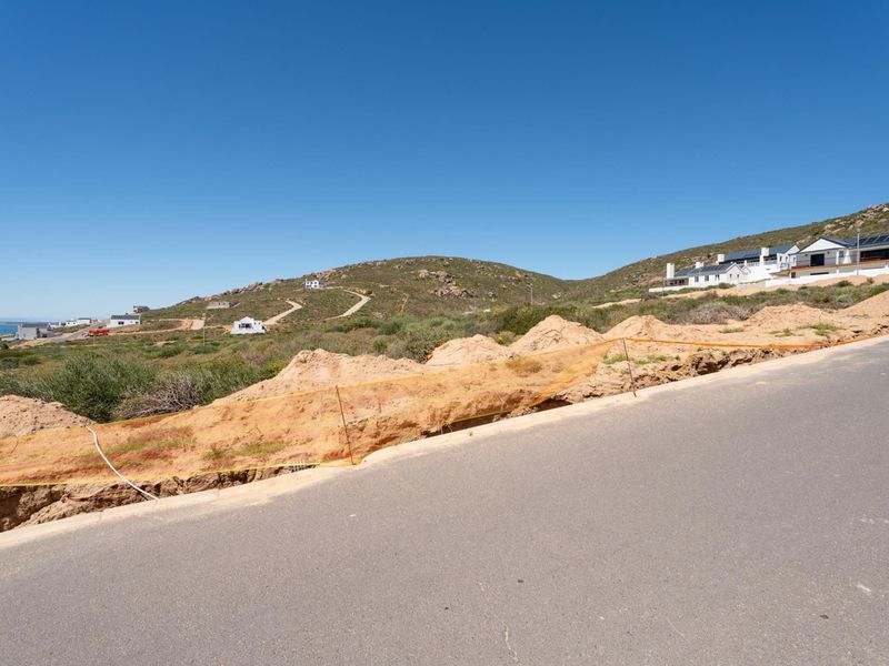 A street-level view of a vacant residential plot featuring an active excavation trench along the paved road in a coastal hillside development.